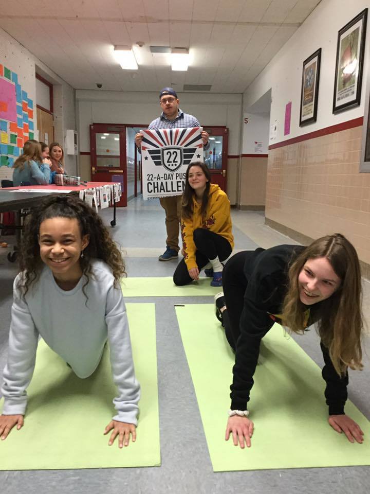 Three students do push ups on yoga mats while a four holds a 22 A Day sign