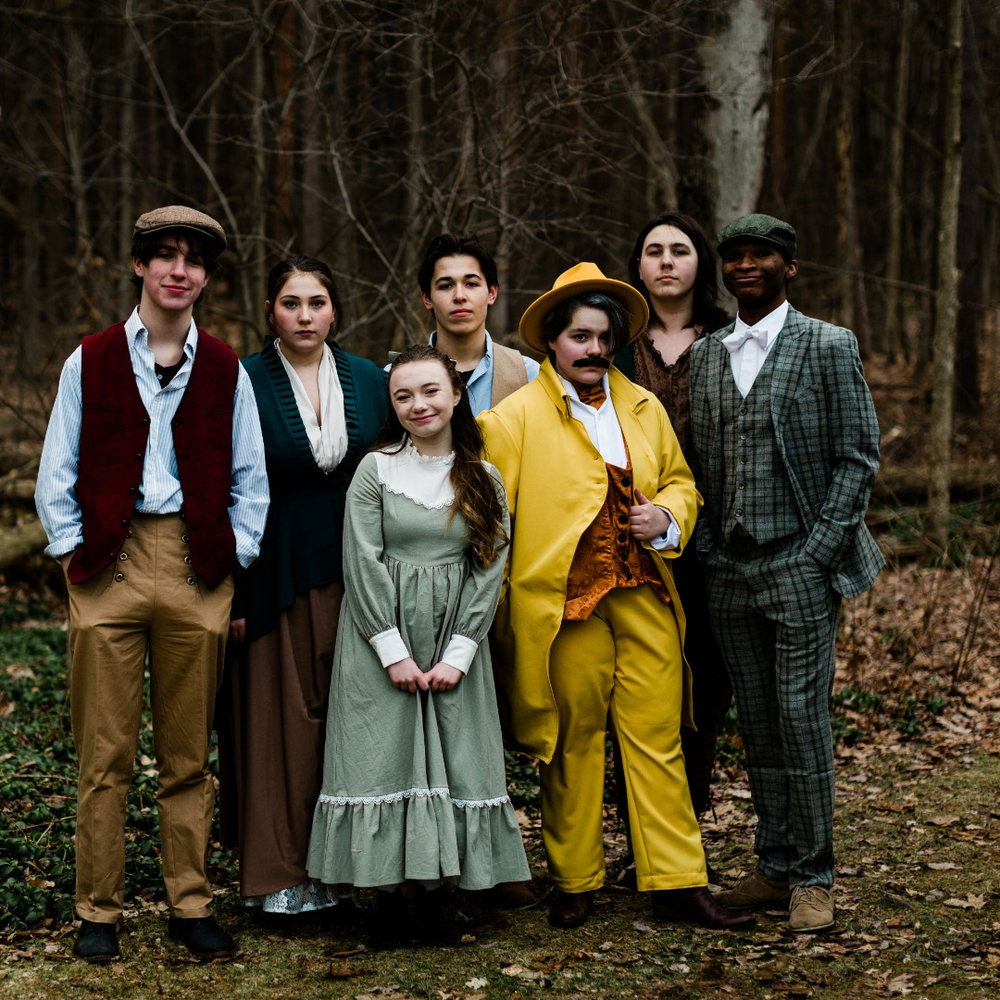 Seven members of the Tuck Everlasting cast stand in a wooded area in their costumes